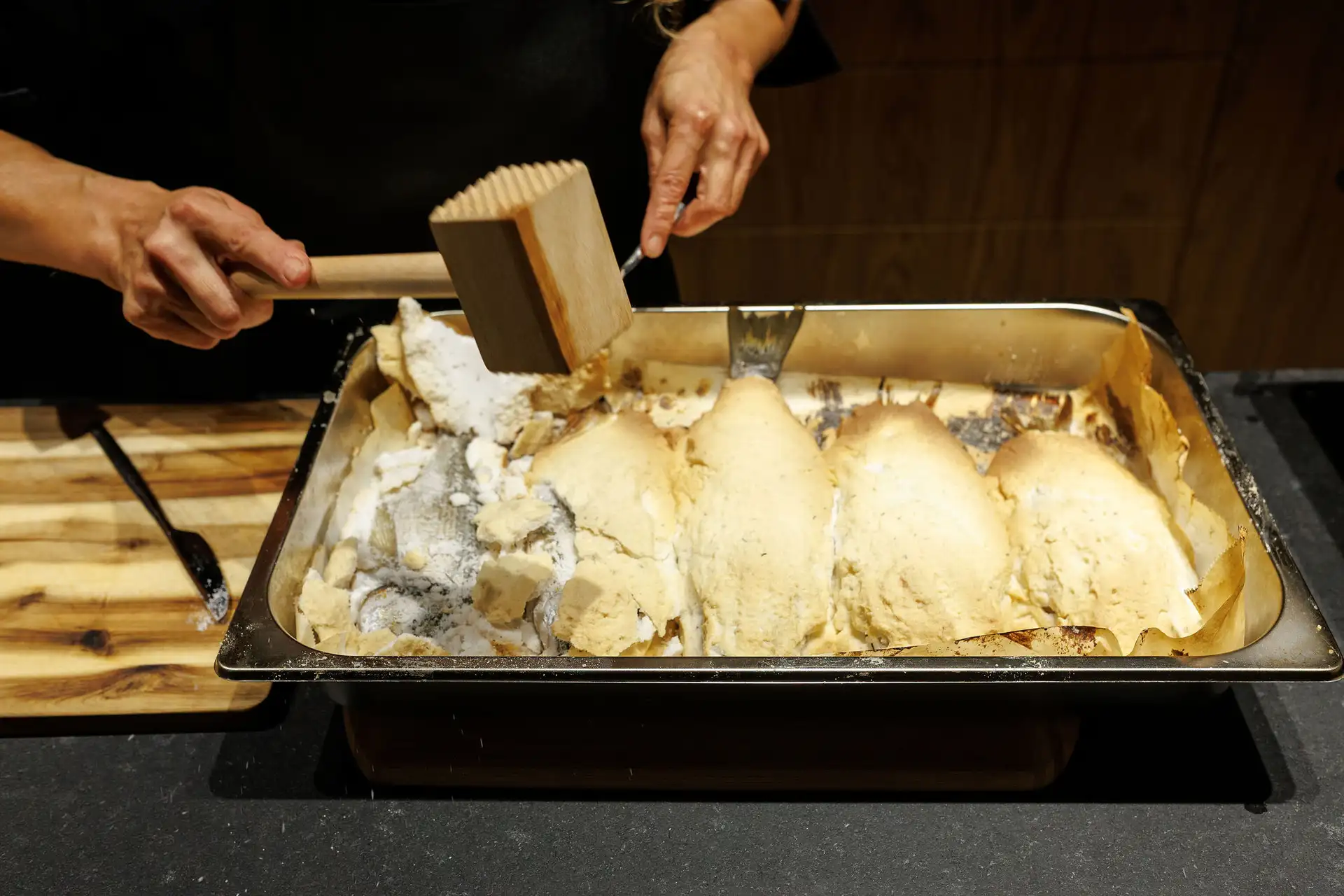 A private chef using a wooden mallet to crack open a salt crust on baked whole fish in a luxury villa kitchen.