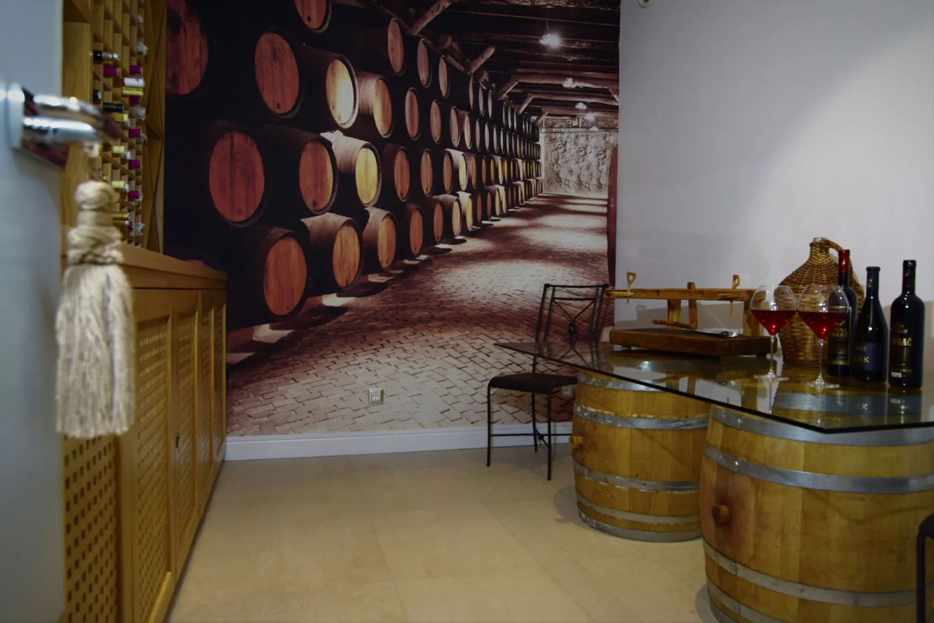 A private luxury villa wine cellar featuring wooden barrels holding up a glass tasting table with red wine glasses and bottles against a vineyard mural backdrop