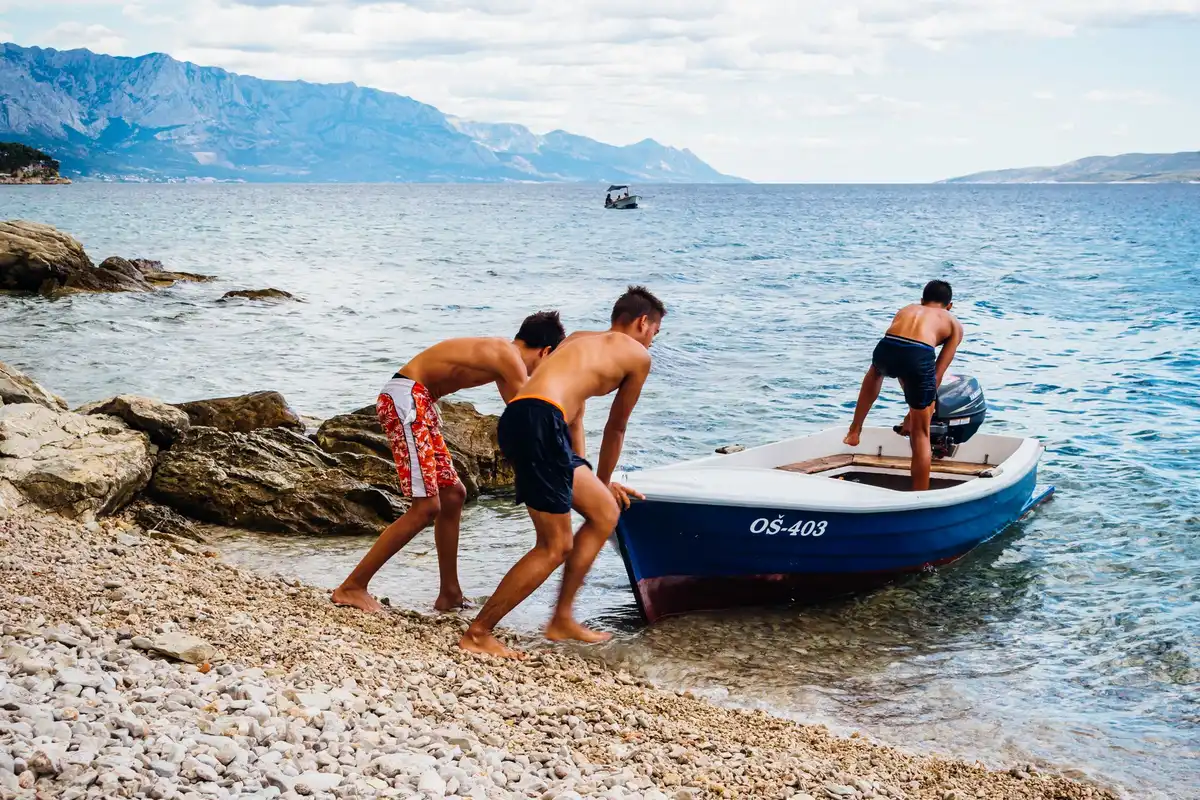 Teenagers pushing a small boat into the sea on the Croatian coast, with mountains in the background
