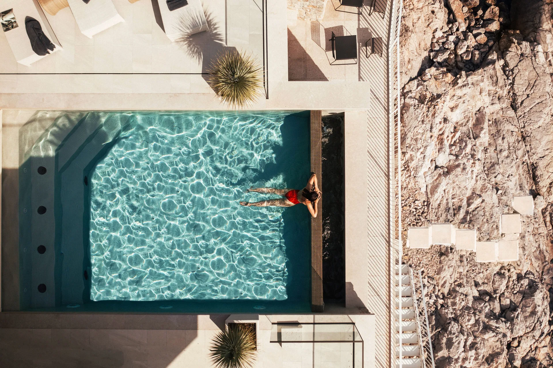 Top-down view of a luxury villa infinity pool with turquoise water next to a rocky coast, featuring a woman in a red swimsuit relaxing at the edge.