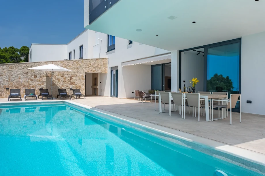A spacious swimming pool accompanied by a large relaxation area with tables, chairs, deckchairs, and an umbrella