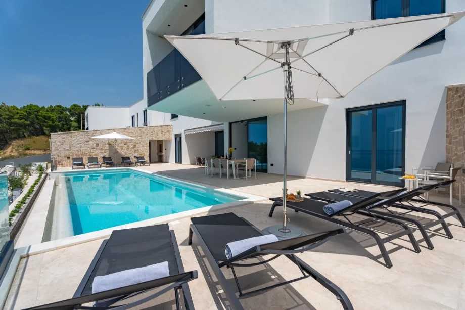 A spacious swimming pool accompanied by a large relaxation area with tables, chairs, deckchairs, and an umbrella