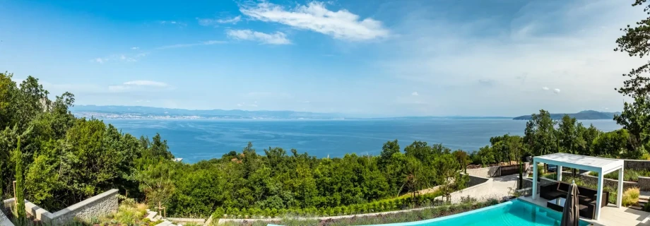Panoramic view of a serene landscape from Villa Aura with a sparkling blue pool in the foreground, lush green trees, and a Adriatic sea under a clear blue sky.