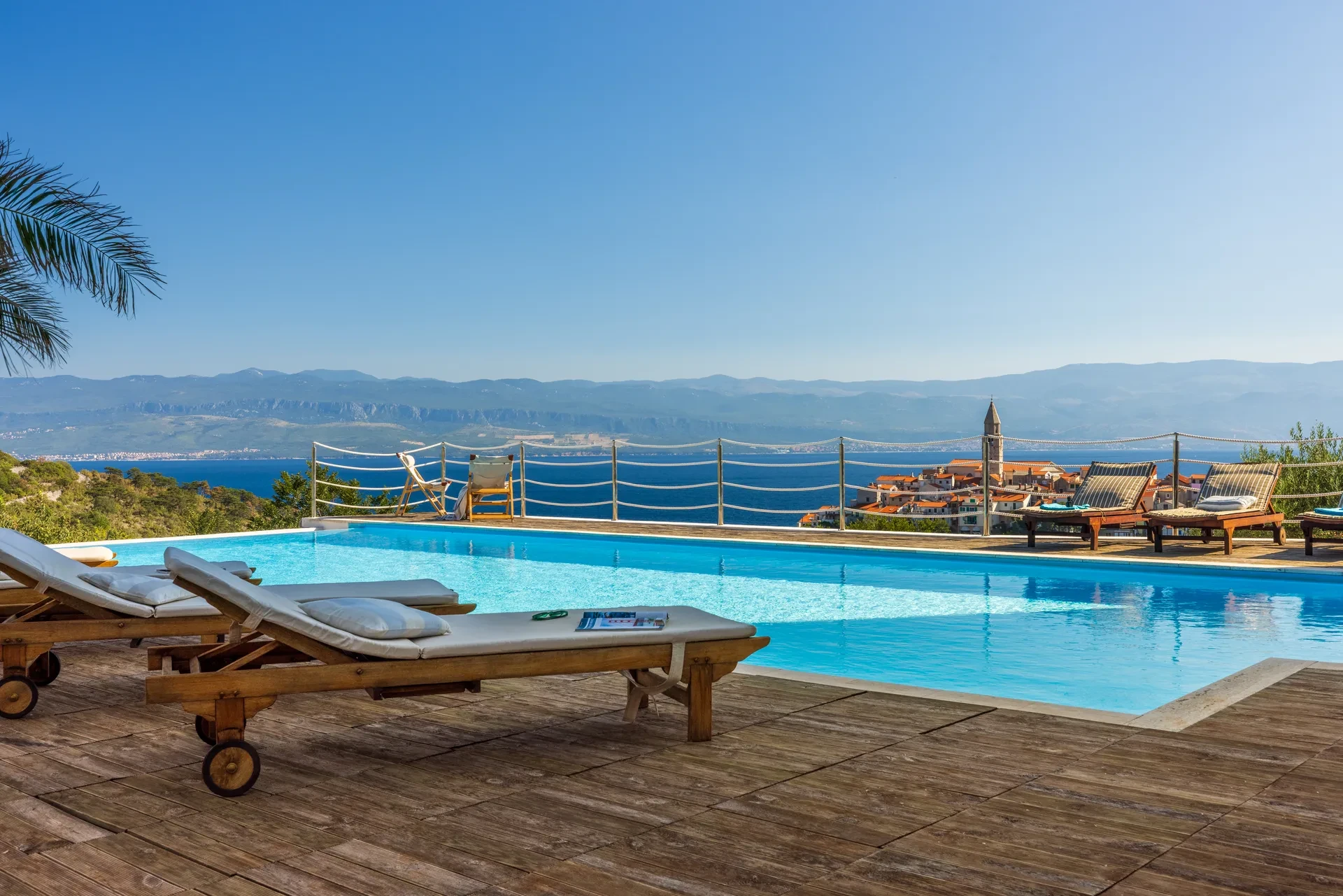 A poolside area with sun loungers at villa Verbanicum overlooks a scenic view of the ocean and mountains on the serene island of Krk under a clear blue sky.