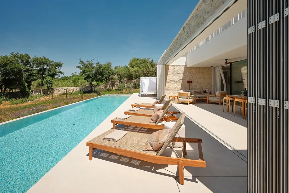 Long private heated swimming pool with sun loungers beside a shaded outdoor living and dining terrace.