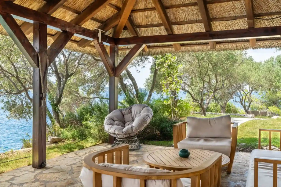Shaded seaside pavilion with lounge seating surrounded by Mediterranean greenery.