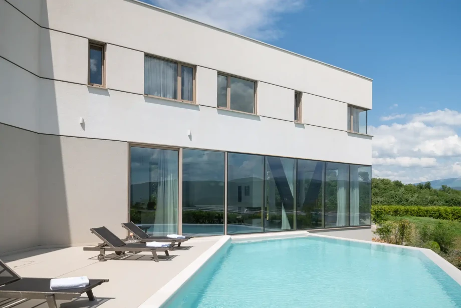 Modern villa with floor-to-ceiling glass opening directly onto the pool terrace and sun loungers